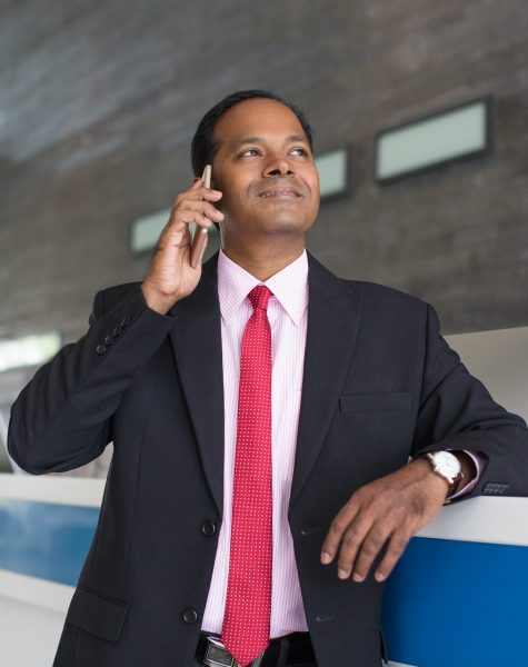 Closeup portrait of content middle-aged Indian business man looking away, calling on mobile phone and standing at hotel reception