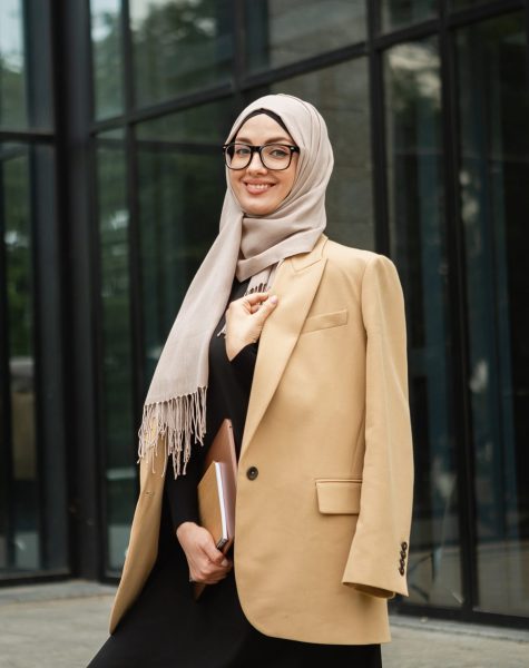 modern stylish muslim woman in hijab, business style jacket and black abaya walking in city street with laptop, freelancer online remote work, business woman in glasses